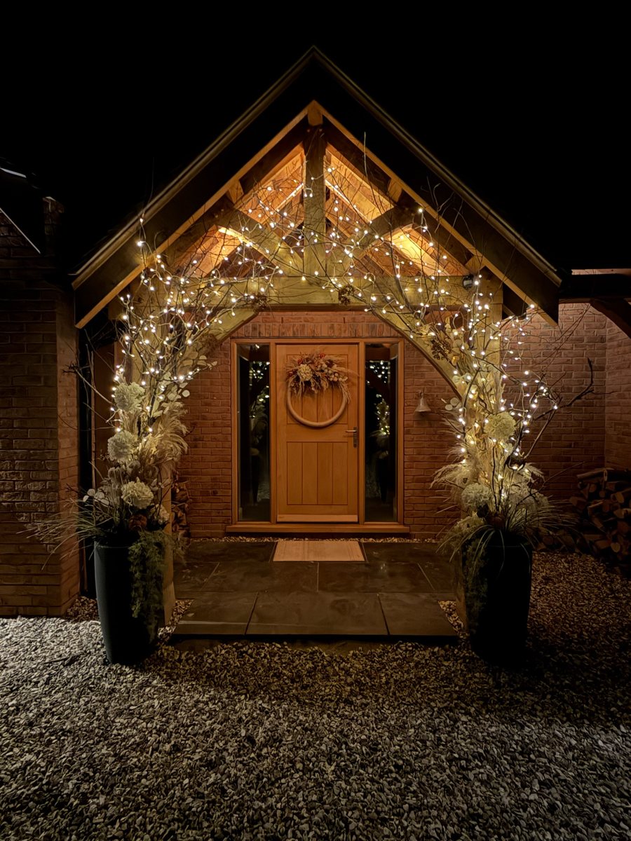 Decorated timber porch at night, with warm fairy lights around the doorway and two tall floral planters.