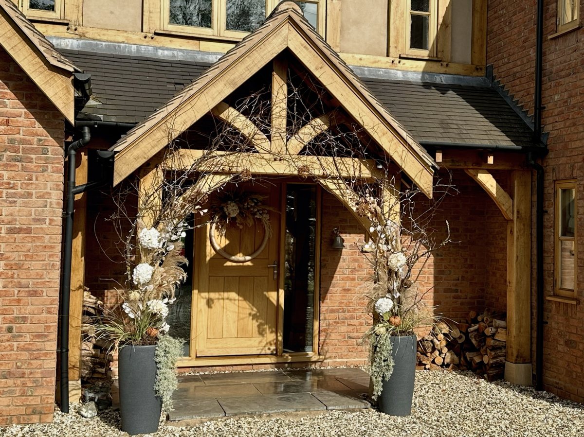 Timber porch framed by branches, cream flowers and greenery in two tall planters.